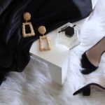 Affordable Luxury - pair of gold-colored earrings on table and black ankle-strap pumps on area rug